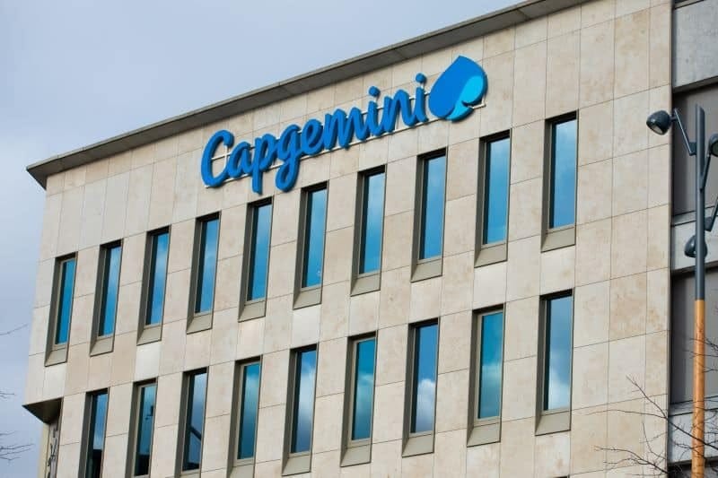 Bulutlu gökyüzü altında, açık renk taş kaplamalı ve sıra sıra pencereli bir ofis binasının cephesinde mavi Capgemini logosu.