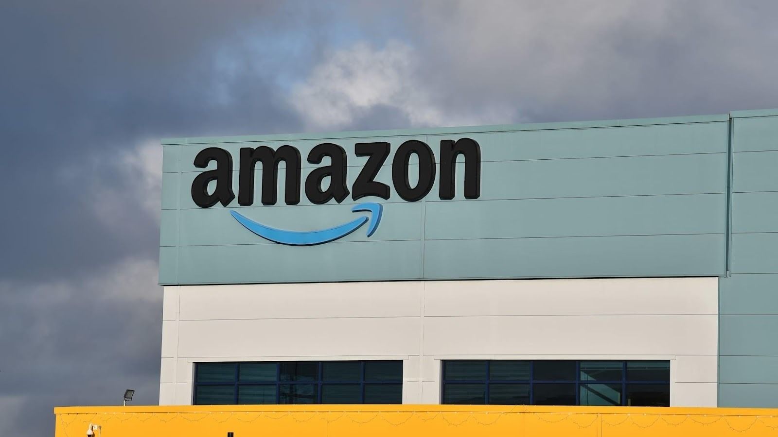 Bulutlu gökyüzü önünde, yeşilimsi mavi cepheli bir binanın üst kısmında büyük Amazon logosu.