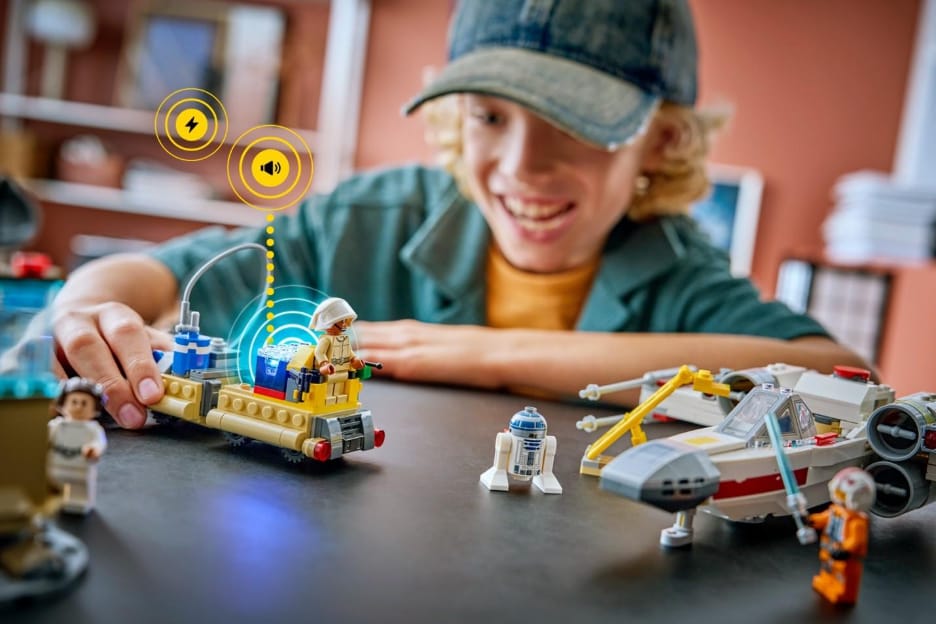 Gülümseyen bir genç, masadaki LEGO Star Wars aracını eliyle sürüyor; araçtan yükselen ses ve ışık simgeleri Smart Brick’in tepkisini gösteriyor, önde R2-D2 ve X-Wing modelleri duruyor.