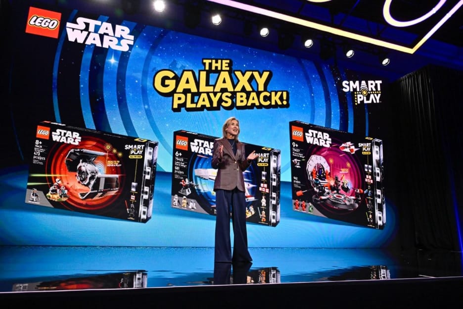 CES sahnesinde bir sunucu, arkasındaki dev ekranda “LEGO Star Wars – The Galaxy Plays Back!” yazısı ve Smart Play logolu üç Star Wars setinin kutusu.