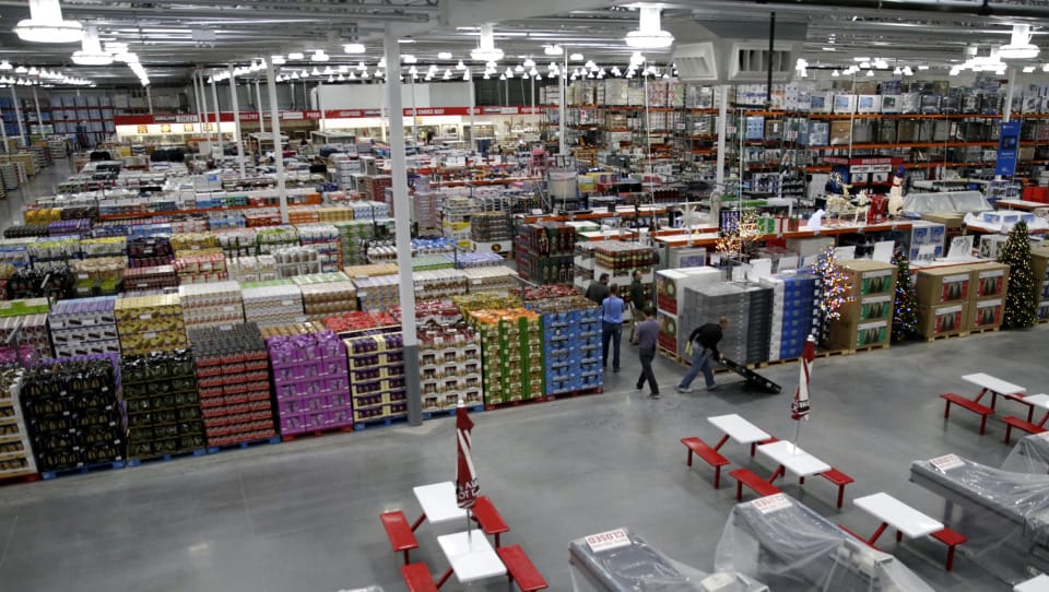 Yukarıdan görülen geniş bir Costco mağazasının içi: palet palet istiflenmiş ürünler, yüksek raflar, alışveriş yapan müşteriler ve kırmızı-beyaz piknik masaları.