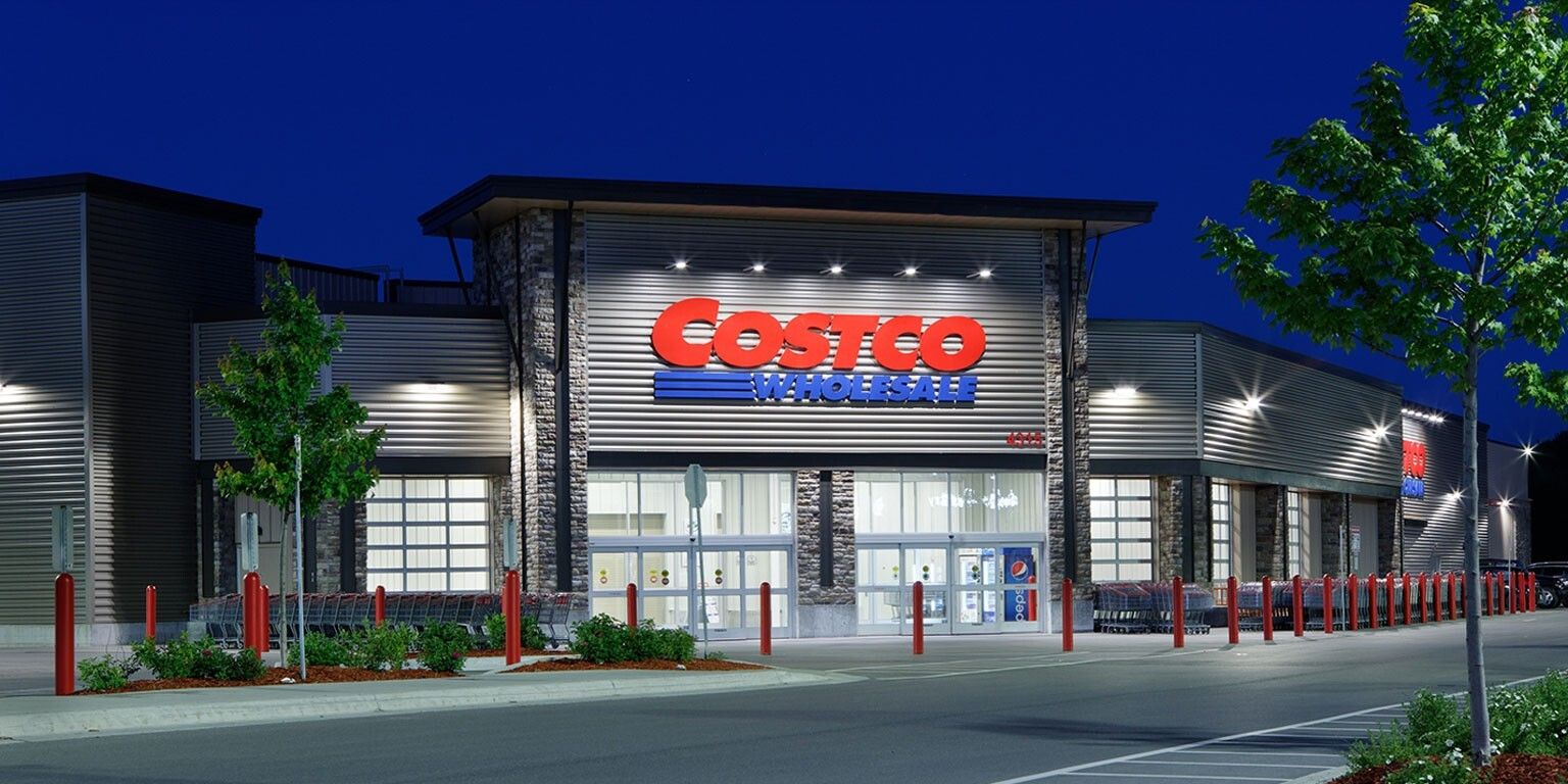 Gece lacivert gökyüzü altında ışıklandırılmış bir Costco Wholesale mağazasının cephesi, cam girişleri ve önündeki kırmızı koruma direkleri.