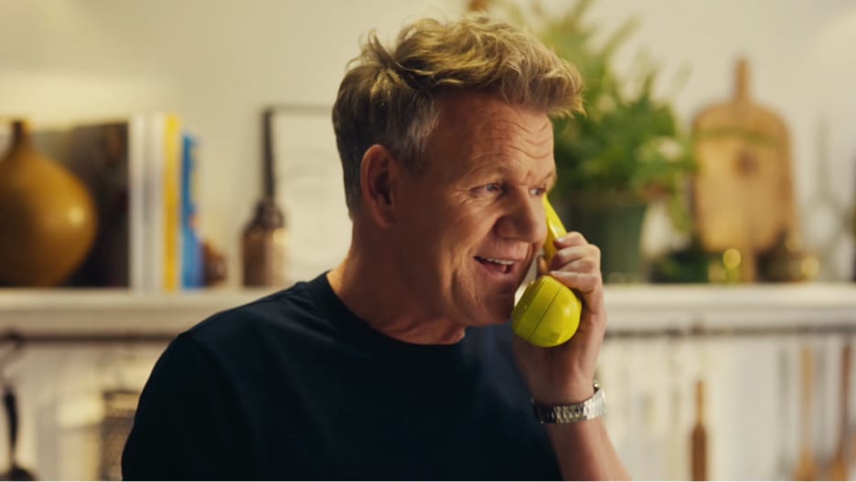 PolyAI reklam filminden bir kare: siyah tişörtlü Gordon Ramsay, raflı bir mutfakta sarı bir telefon ahizesiyle konuşurken gülümsüyor.