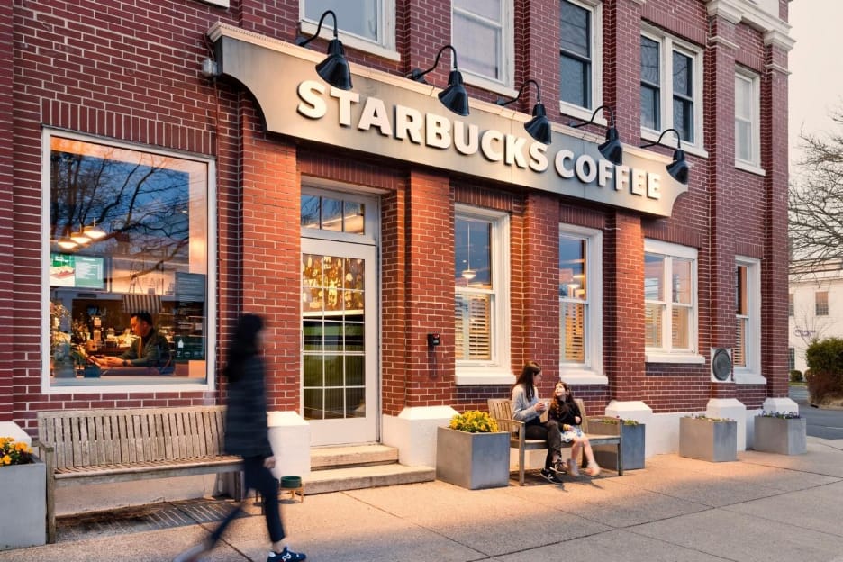 Kırmızı tuğlalı bir binadaki Starbucks Coffee mağazasının akşam vakti cephesi; önündeki bankta oturan iki kişi ve kaldırımdan geçen bir yaya.