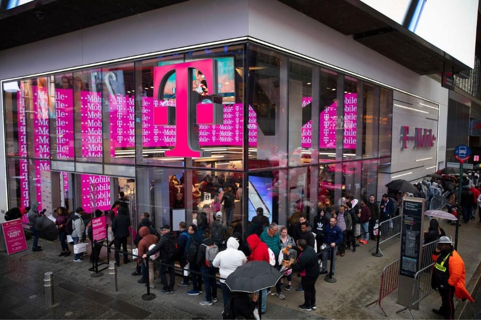 Pembe T-Mobile logolarıyla kaplı camlı bir mağazanın önünde, yağmurlu bir günde şemsiyeli ve montlu insanların bariyerler boyunca kuyruk oluşturduğu kalabalık